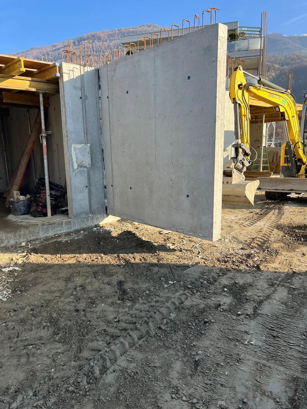 Voile de béton décoffré devant une pelle jaune et les montagnes